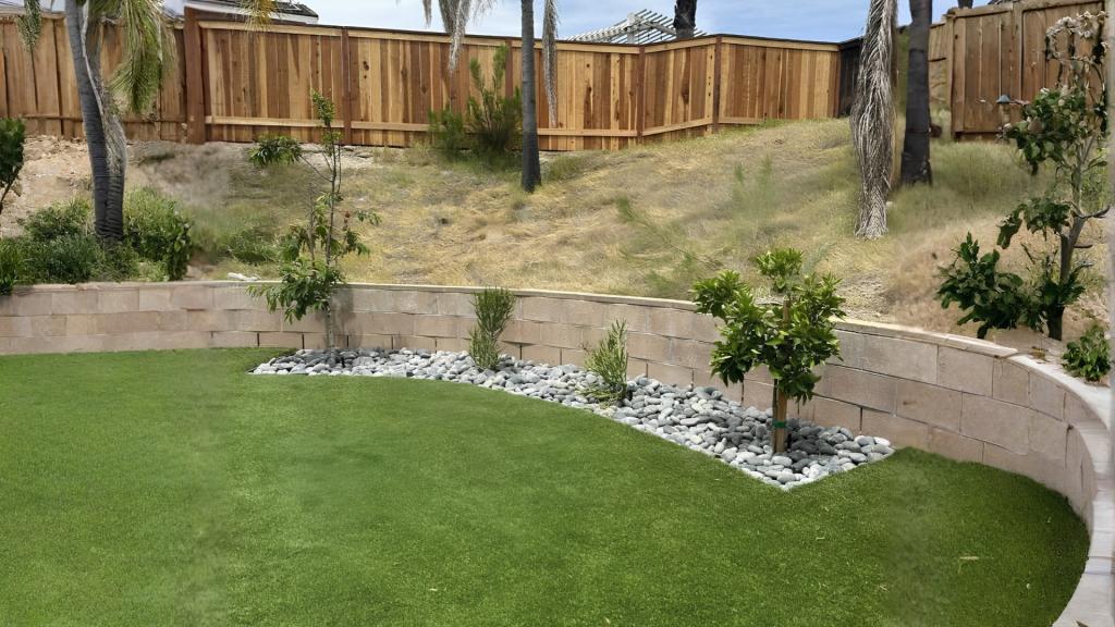 Backyard landscaping in San Diego with artificial turf, curved retaining wall, river rocks, young trees, and a wooden privacy fence